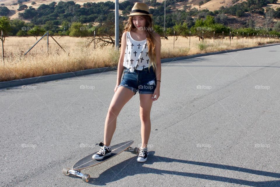 Teenage girl in shorts and converse shoes with one foot on a longboard on the street with a field and hills in the background
