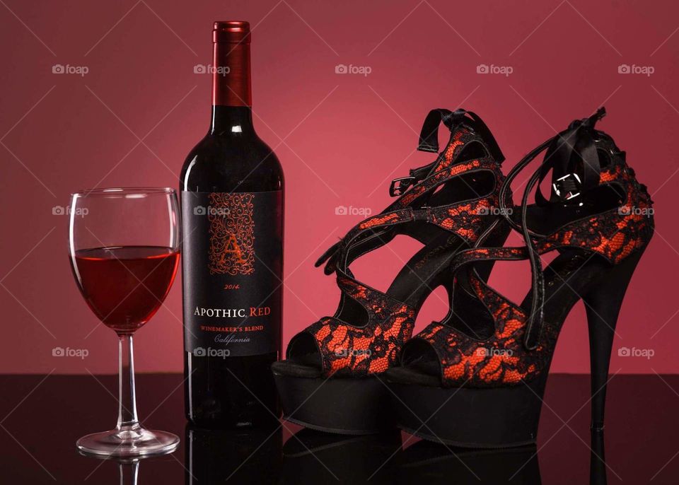 red wine in a glass next to a black bottle of red wine with red labels next to a pair of red shoes with black lace accents all in the foreground with a red gradient background