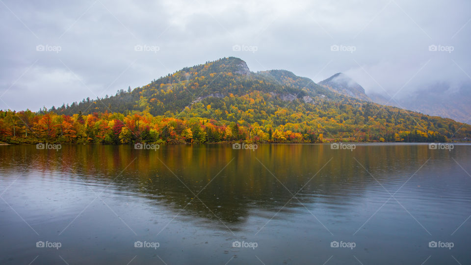 Echo Lake in rain, with clouds and mist lingering with mountains in morning. Reflection in water of colorful yellow, red and green autumn leaves. Franconia Notch, NH, USA. Background of autumn leaves. Rainy weather. New England fall foliage.