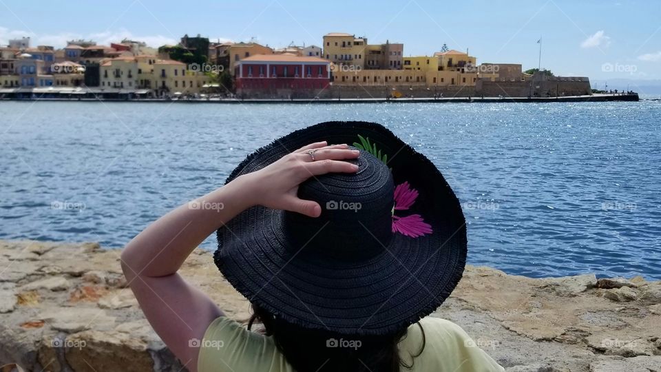 Woman holding hat sitting on bench looking out to ocean and beautiful buildings on sunny summer vacation