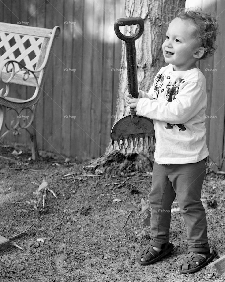 A cute little toddler boy plays with a toy garden rake in the backyard on a sunny summer day. 