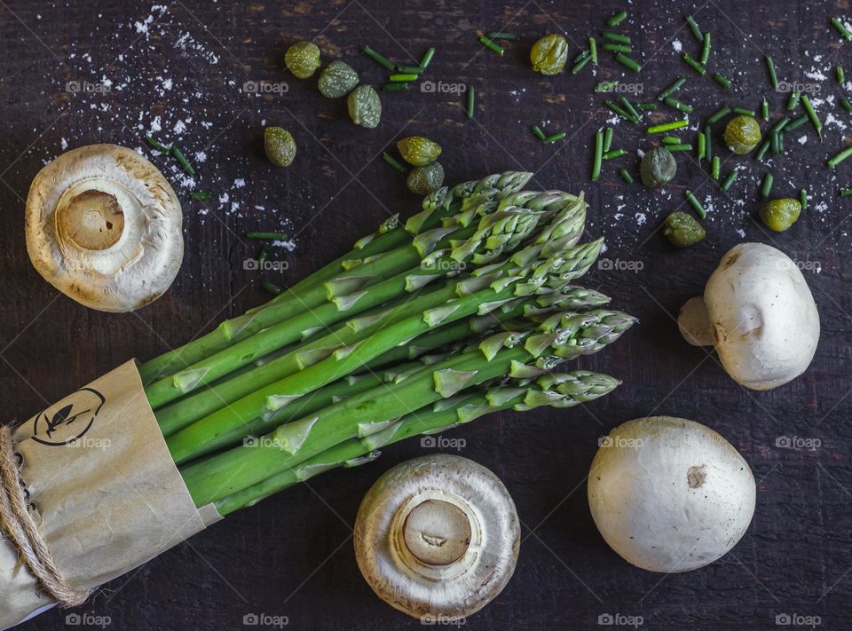 Asparagus themed flat lay with mushrooms, capers and chives on a dark wood background 