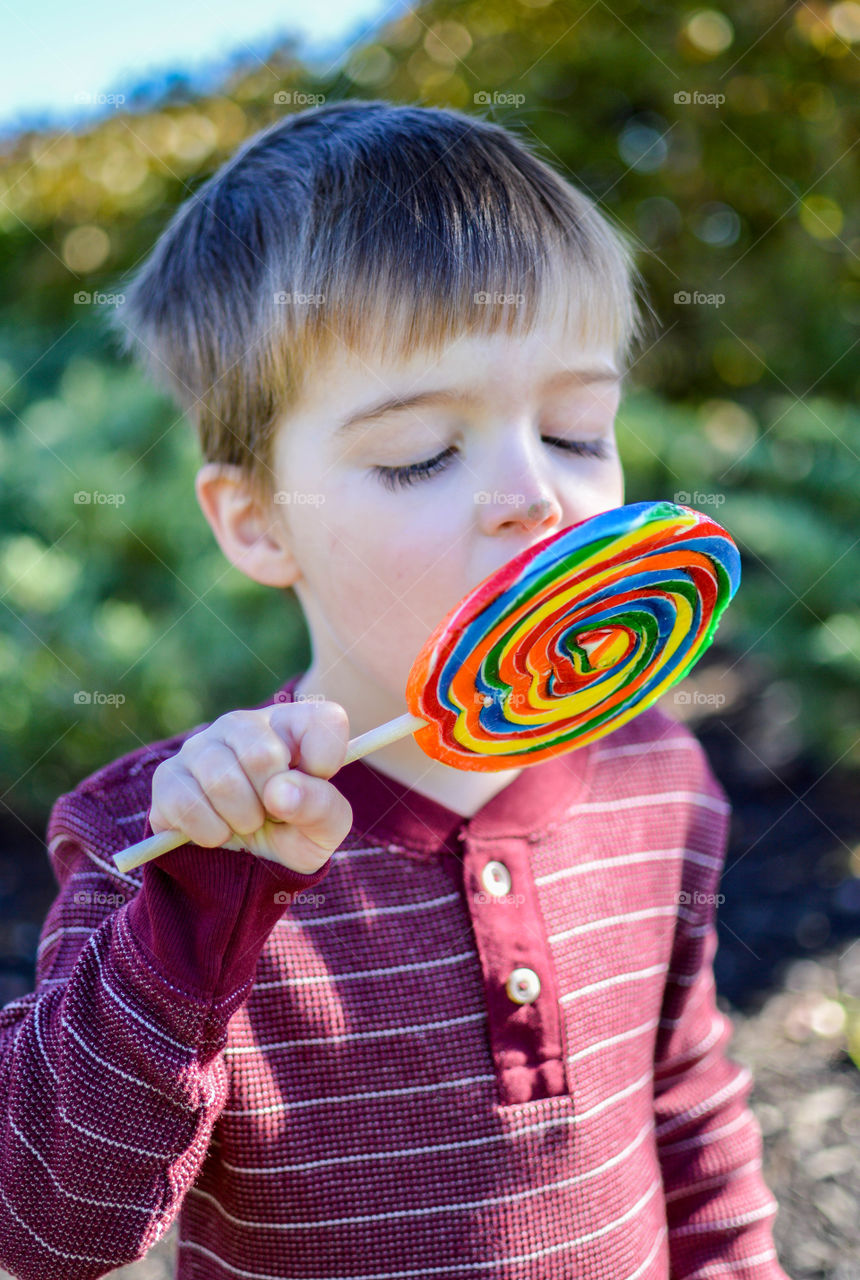 Young boy eating a large rainbow colored lollipop outdoors