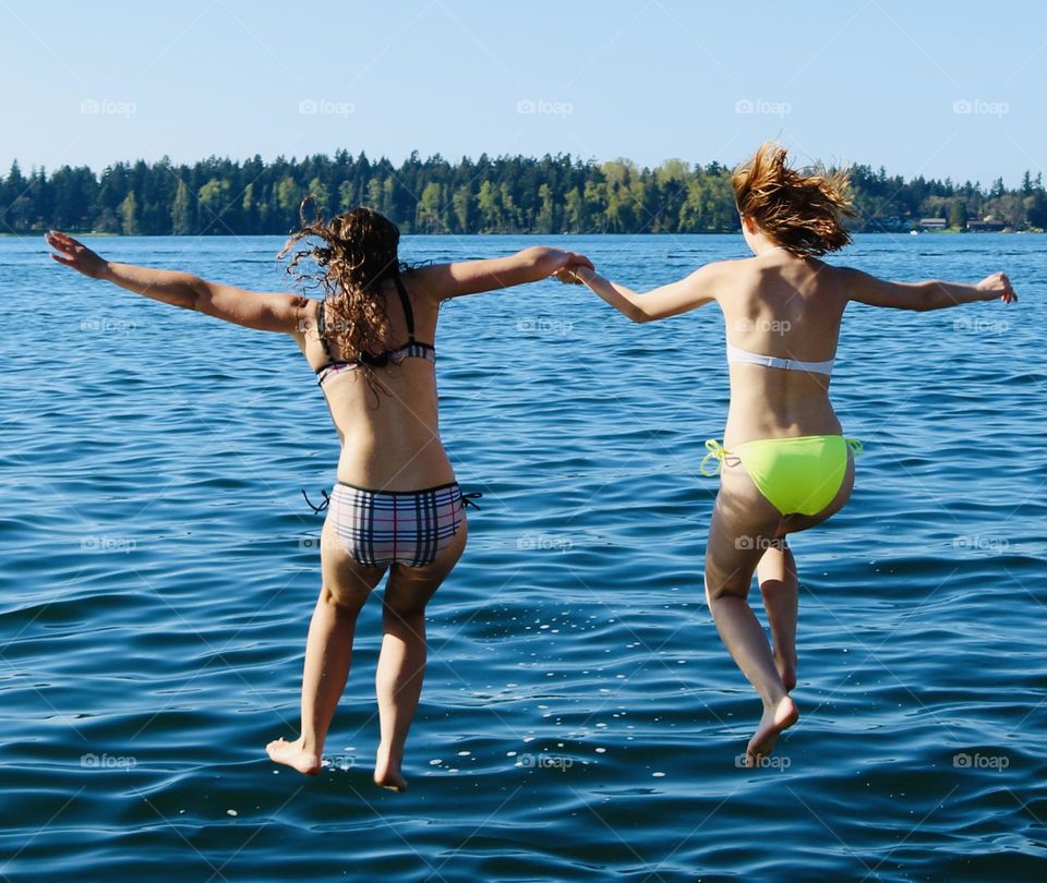 Two young women in bikinis join hands as they jump into the refreshing blue water of American Lake, Washington 