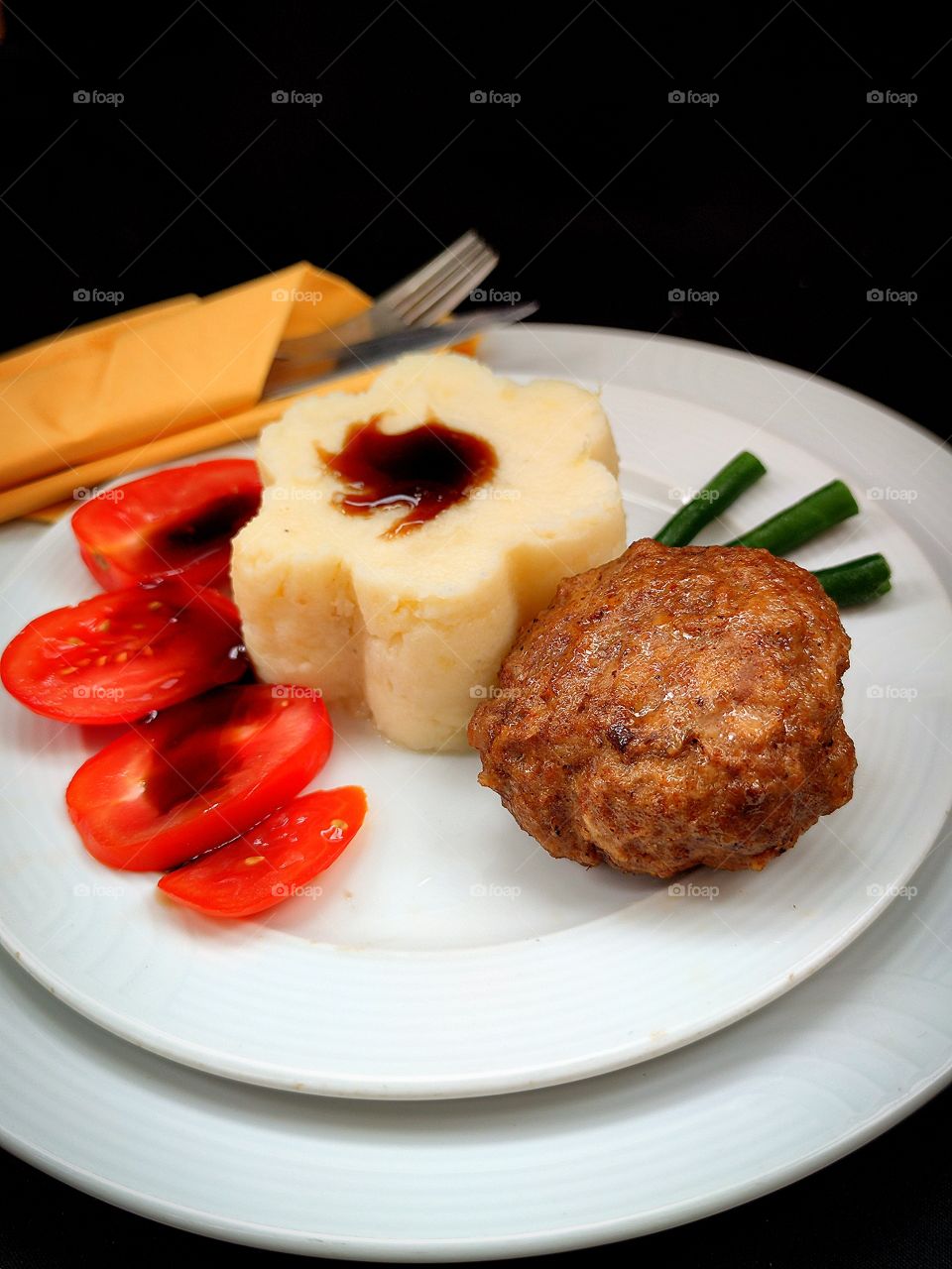 On a white plate are: meat cutlet, flower-shaped mashed potatoes, chopped tomatoes and three stitches of green beans.  On the bottom plate are a fork and a knife, which are wrapped in a napkin